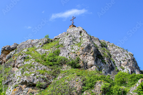 A tall cross on a rock covered with bushes and grass. Blue sky with clouds. Elia, Kakheti, Georgia