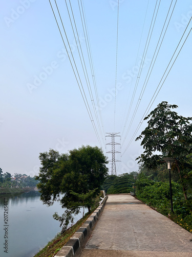 Walkway running alongside a calm lake with lush trees and landscaped greenery beneath overhead high-voltage transmission lines.
