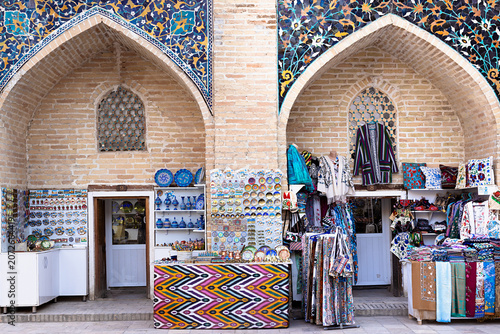 Souvenir shops inside Nadir Divan Begi Madrasah in Bukhara displaying colorful ceramics and textiles beneath ornate tiled arches. Ideal for travel publications and cultural tourism content.