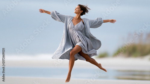 Joyful woman leaping along windswept beach in flowing dress and cardigan