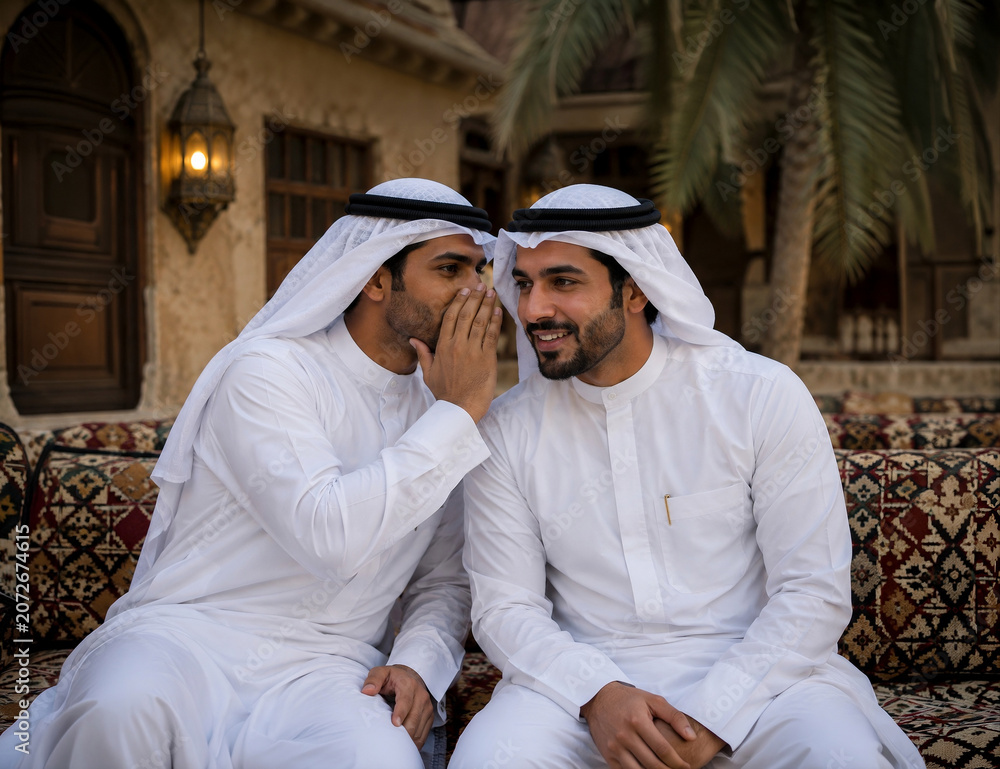 Fototapeta premium Arab Man Whispering a Secret to Friend in Traditional Gulf Clothing | Two Arab men wearing traditional Gulf attire sharing a private conversation while sitting outdoors