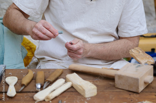 Artisan carving wood by hand at a workbench with traditional tools and wooden pieces in progress.