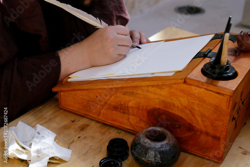 Person writing with a quill on parchment sheets at a traditional wooden writing desk.