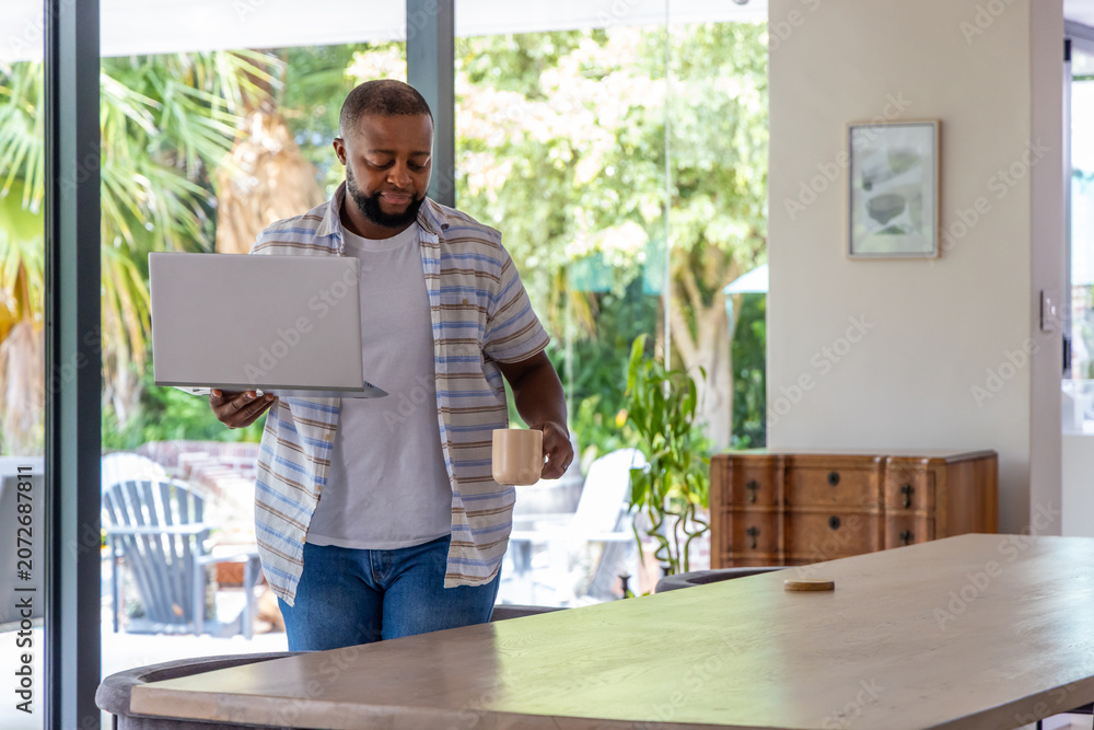 Fototapeta premium African American man standing in dining area holding open laptop and ceramic mug near doors