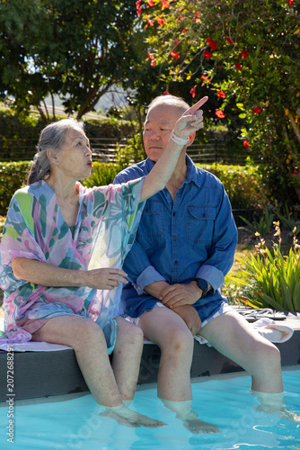 Diverse senior couple wearing pastel cover-up and denim sitting on pool edge pointing at red blooms