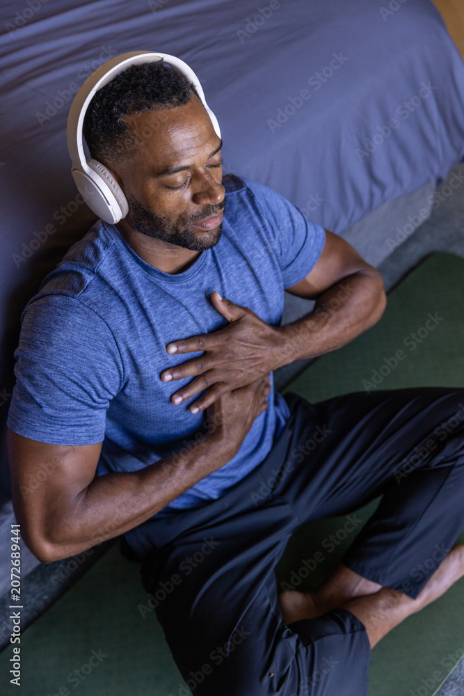 Fototapeta premium African American man meditating on green mat beside bed, wearing blue shirt and white headphones