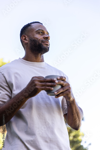 African American man standing in yard holding ceramic cup, wearing light tee, ringed left hand