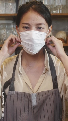 Vertical Screen : Young Asia female restaurant staff wearing protective face mask using infrared thermometer checker temperature gun on customer's forehead before enter. Lifestyle after corona virus.