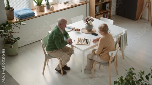 Wide angle shot of senior man and senior woman playing chess at table in retirement home. Clip for marketing campaigns promoting active aging and senior care facilities