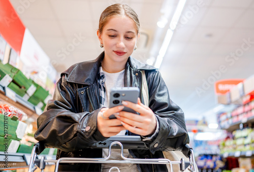 Young Girl Goes Shopping With A Cart In The Supermarket Using A Shopping List On Smartphone