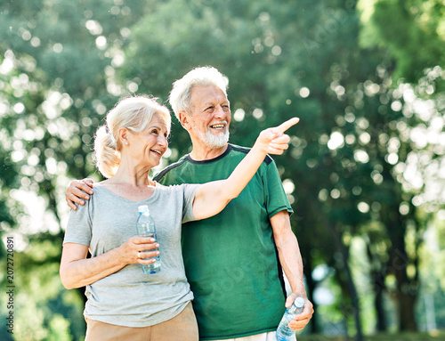 Smiling active senior couple holding water bottles, drinking and  jogging together in the park