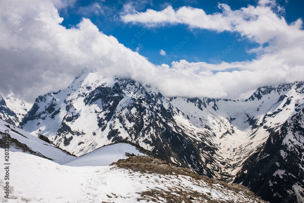 Fototapeta premium Snow-covered mountain range features rugged peaks and a valley, with a clear blue sky and clouds creating a dramatic landscape