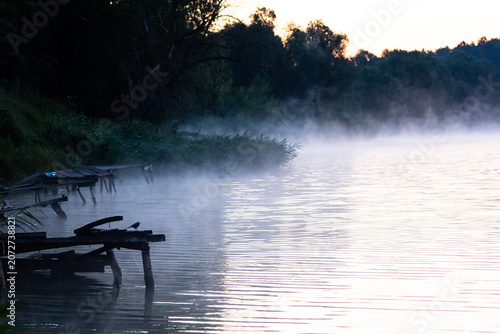 Quadro su tela Thin mist rolls along a reedy bank dotted with old wooden piers while morning li