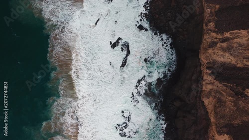 Top down drone view of powerful Atlantic waves breaking against the rocky volcanic coastline of Fuerteventura, Canary Islands, Spain. Turquoise ocean and white sea foam.