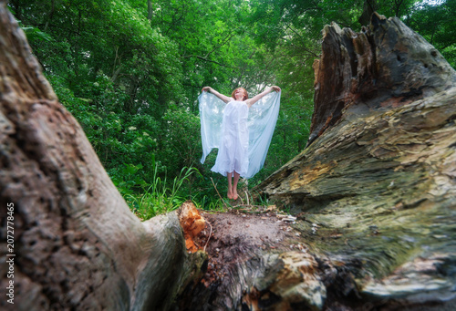 Happy woman jumping in flowing white fabric between ancient tree trunks in a lush forest creating a joyful woodland scene filled with freedom wonder and vitality
