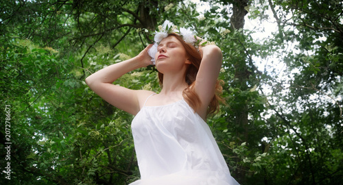 Red haired woman in a flowing white dress wears a floral crown with closed eyes beneath forest trees. The portrait expresses harmony spirituality renewal and quiet imagination
