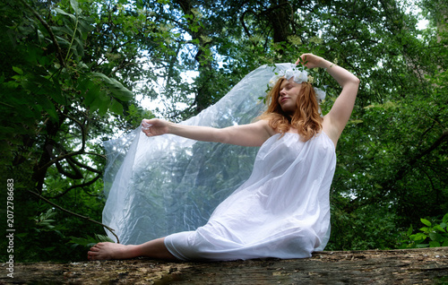 Redhead woman wearing a flowing white dress and floral crown sits on a forest log with translucent fabric. The dreamy portrait evokes mythology transformation freedom and imagination