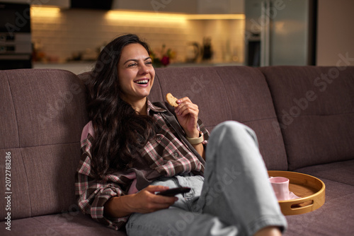 Woman feeling happy, laughing and relaxing on a sofa at home, eating a cookie and watching tv in the evening