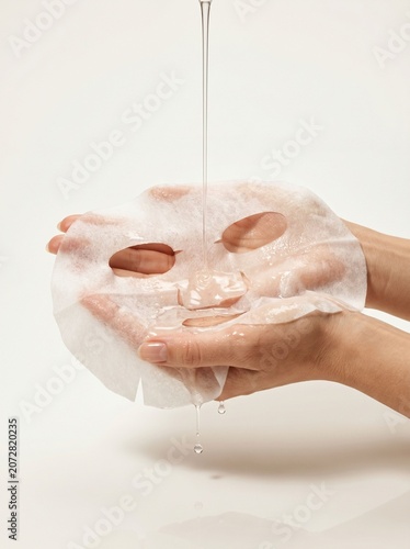 Clear liquid pouring onto white facial sheet mask held by hands
