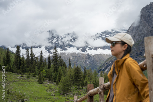 The female traveler stands with her back against a fence, with snow-capped mountains and a forest in the background