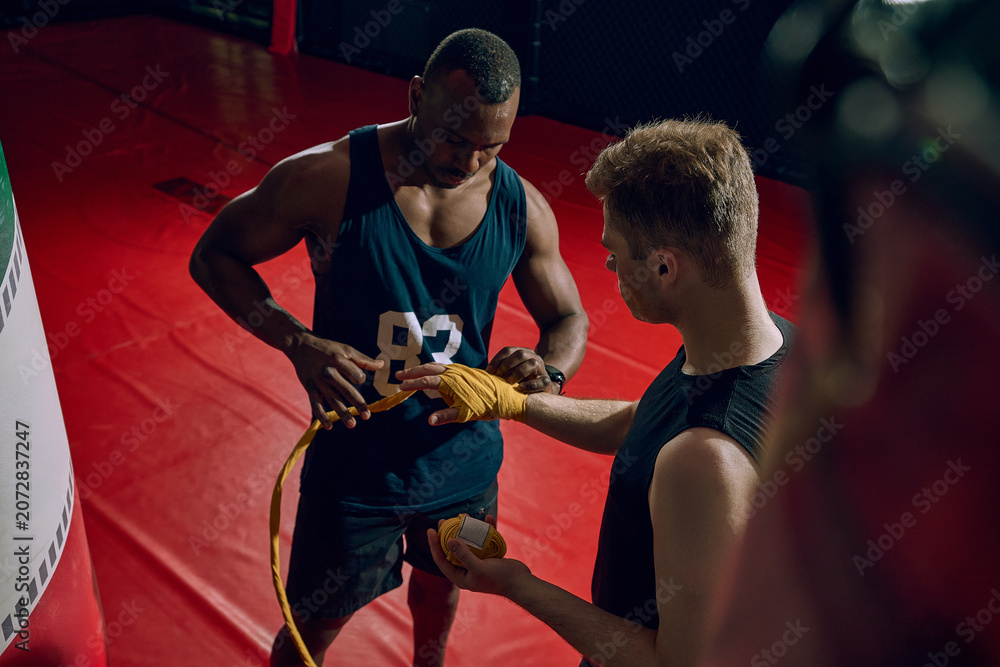 Naklejka premium Boxing coach wrapping athlete hands before training session in dark gym. Concept of safe preparation, coach mentorship, injury prevention, teamwork and sports education.