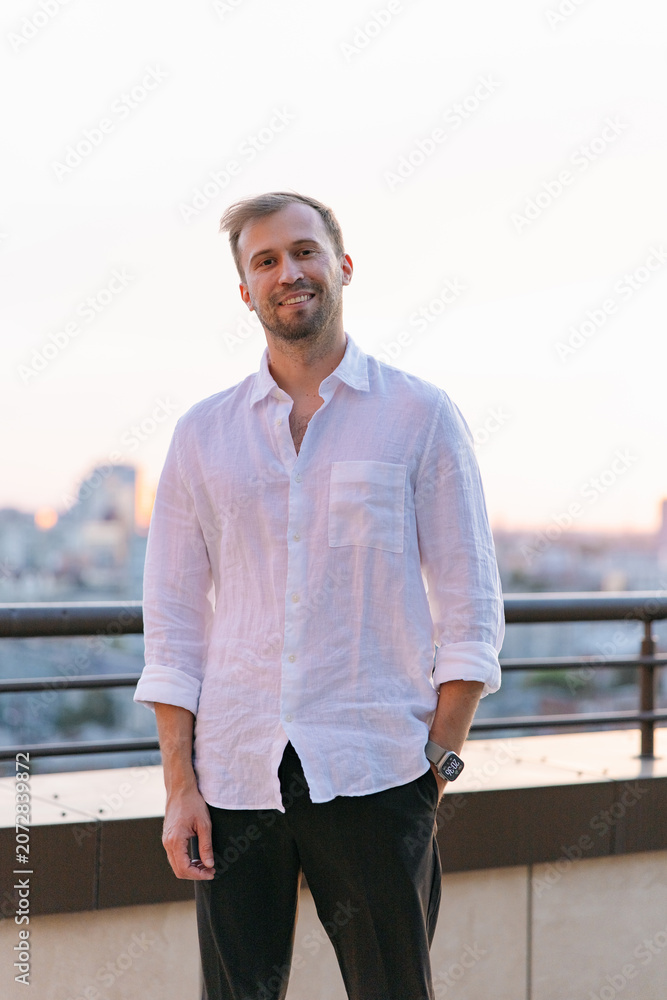 Fototapeta premium Smiling handsome young man in white linen shirt standing on rooftop terrace at sunset