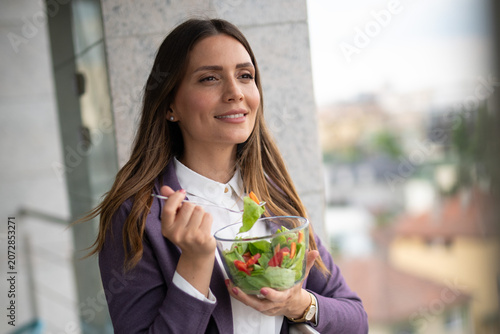 Smiling woman in business attire enjoys a healthy salad outdoors on a balcony