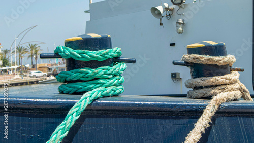 Bright green synthetic and classic hemp mooring ropes tied around metal bitts on a commercial vessel at a port during daytime