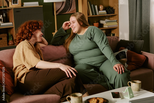 Young plus size women sitting on sofa, talking and laughing during home visit, showing friendship, emotional support, body positivity. Ideal for wellness, community, lifestyle marketing