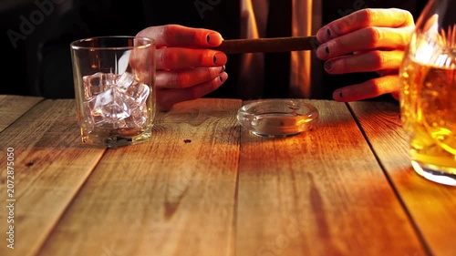 Person holding a cigar while sitting at a table with whiskey glasses and poker chips in a dimly lit setting.