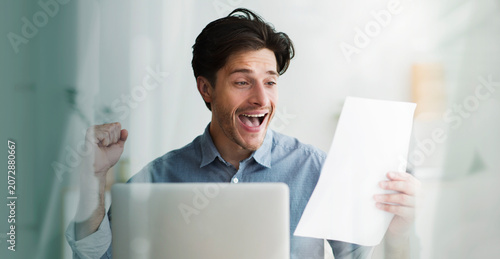 Fotografie Excited Caucasian Man In Casual Attire Smiling And Celebrating Success While Reading Document From Screen In Modern Office Environment