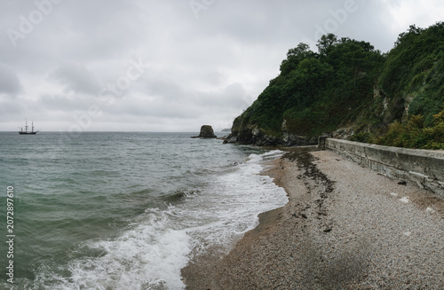 Gravel and rough sand beach near city of Charlestown in Cornwall on overcast day, small classic sail ship on sea distance