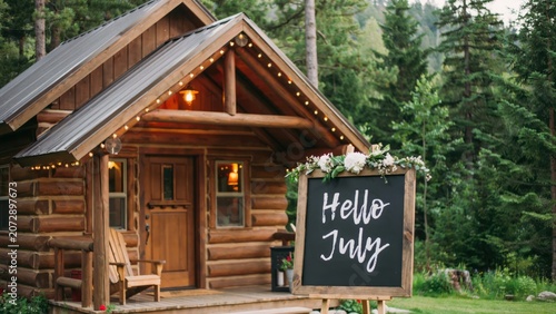 Hello July message on a chalkboard easel outside a cozy rustic log cabin in a green forest