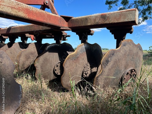 Obraz na plátně Rusty Plow Disc Blades Against Blue Sky Field
Row of wavy rusty plow disc blades on red frame against bright blue sky, agricultural tillage equipment on dry grass field, rural farming detail