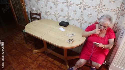  Mujer mayor con bata rosa usando un teléfono inteligente en la silla de la cocina. Mujer mayor con bata rosa y gafas navegando por un teléfono inteligente sentada en la cocina. Senior activo, rutina 