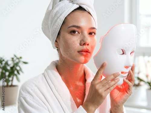 Young woman in bathrobe holding a white led light therapy mask
