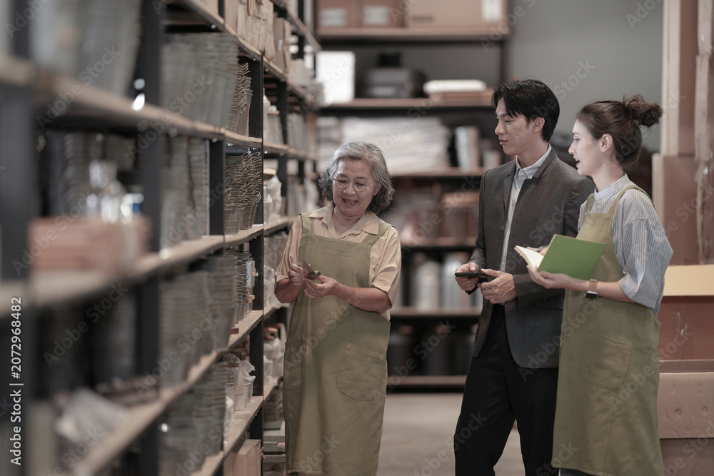 Fototapeta premium A young Asian craft shop owner in a product storeroom discussing inventory together in a dark wood-shelved storeroom in a small business warehouse. A picture of small business success.