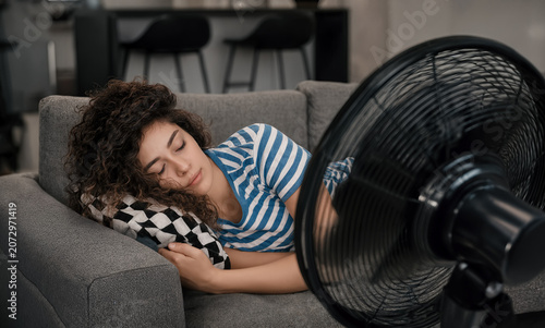 An exhausted woman naps on a couch, facing a powerful electric fan trying to beat the intense heat in a modern room.