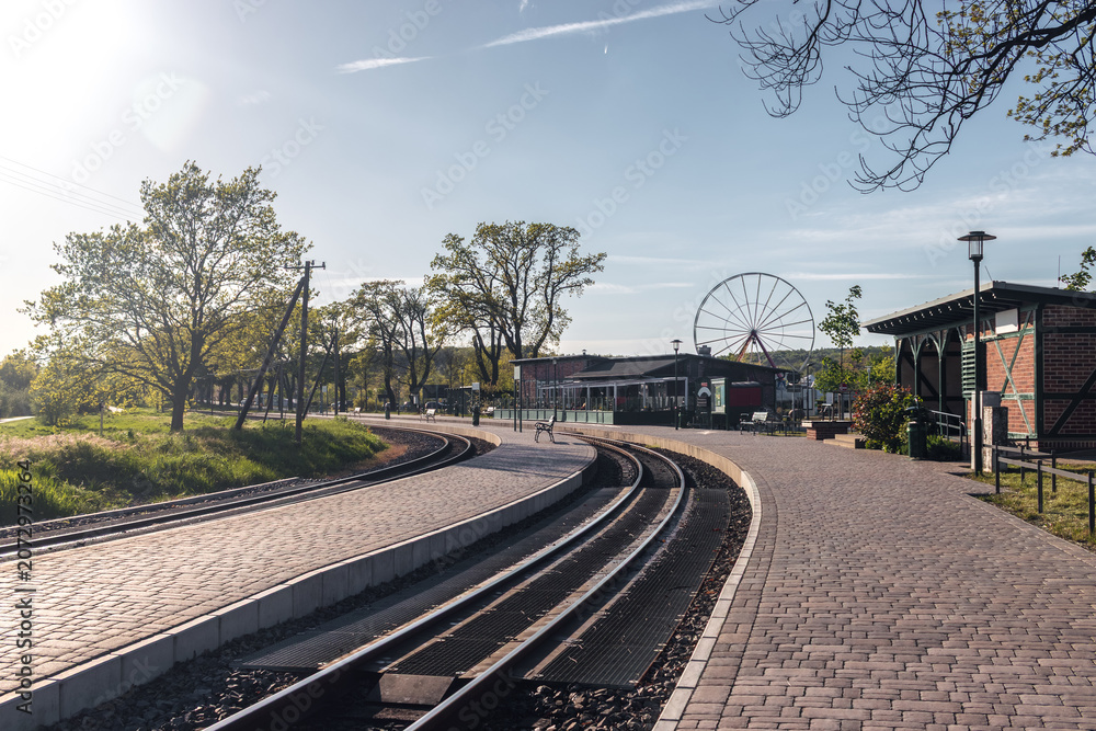 Fototapeta premium View of the narrow-gauge railway station platform and train tracks with a Ferris wheel visible in the background in Sellin, Ruegen island, Germany.