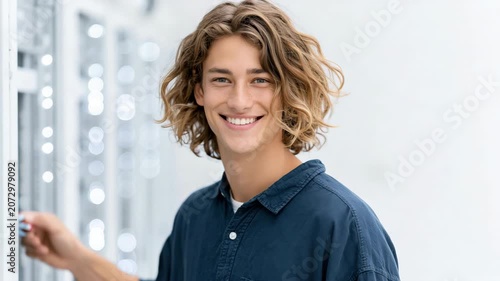 Tracking shot of young man turning and smiling while reaching for metallic panel in bright white studio hallway