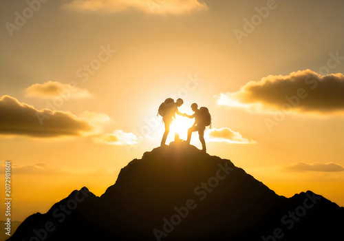 Two hikers helping each other climb to the top of a mountain peak during a beautiful golden sunset