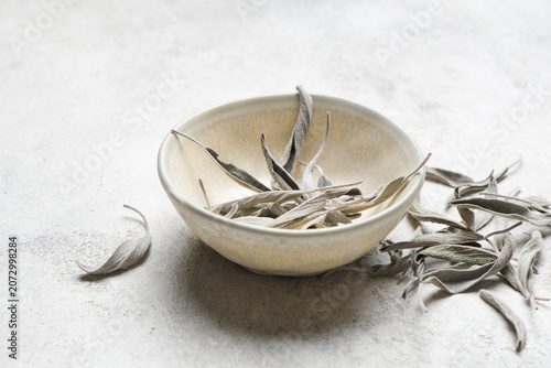 Dry sage herb leaves are placed in a shallow ceramic bowl on a textured gray surface, with additional scattered leaves enhancing the visual arrangement