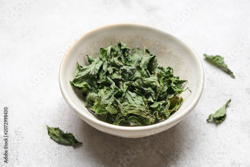 Dried mint leaves arranged in a shallow bowl on a textured surface, highlighting the organic nature and vibrant green color of the herbs