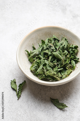 Dried mint leaves are displayed in a round ceramic bowl on a light textured surface, with additional loose leaves scattered nearby