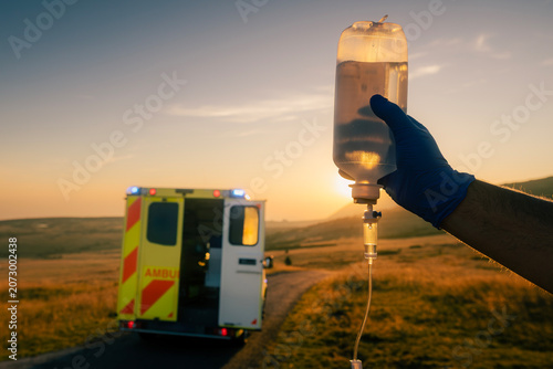 Close-up of paramedic hand holding IV drip on mountain road at sunrise. Emergency medical service, rescue, critical care and rural healthcare concept.