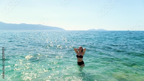 Woman standing waist-deep in turquoise sea, hands behind her head and face tilted toward the sun, handheld summer shot.

