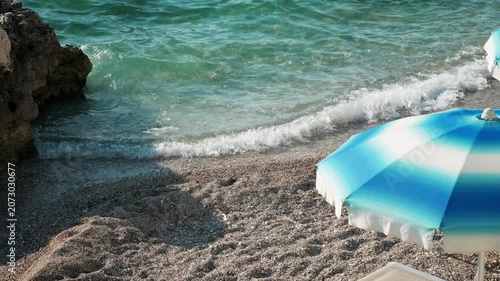 Clear turquoise sea water washing over a sunny pebble beach with a blue and white umbrella by the shore, handheld travel footage of a peaceful summer coastline.