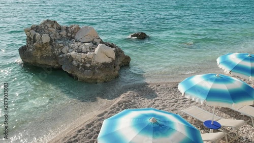 Wide handheld shot of blue and white beach umbrellas, sun loungers and a coastal rock formation on a sunny pebble beach beside clear turquoise sea water.