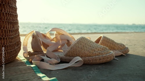 Close up handheld shot of espadrilles on a beach mat with sea in the background, warm sunlight and a peaceful summer travel lifestyle atmosphere.