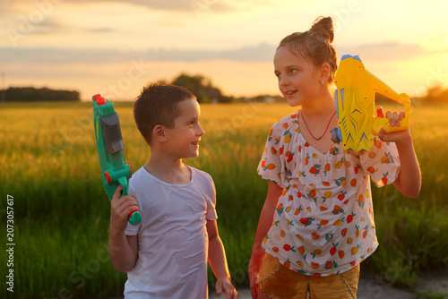 Two children are having fun with water guns in a green field at sunset. They look happy while enjoying their time outside in warm weather during summer